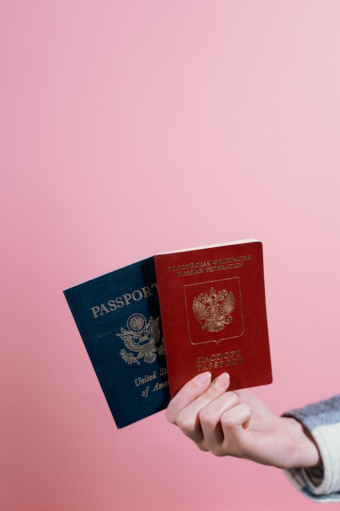 Close-up of two passports held by a hand against a pink background.